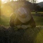 Tired, worried ranch farmer rubbing neck leaning against hay. Backlit at sunset with lens flare