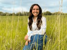 Rebecca Zanello kneels in a field under blue sky. Zanello's PhD work examines livestock risk management tools and grassland conversion decisions for Canadian cattle producers.